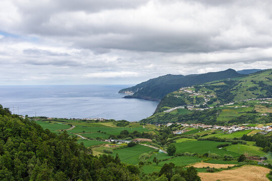 P&ocirc;r-do-Sol viewpoint on S&atilde;o Miguel island showing coastline with lush green fields, rolling hills and charming villages nestled along the Atlantic Ocean under a cloudy sky. Tranquil rural landscape