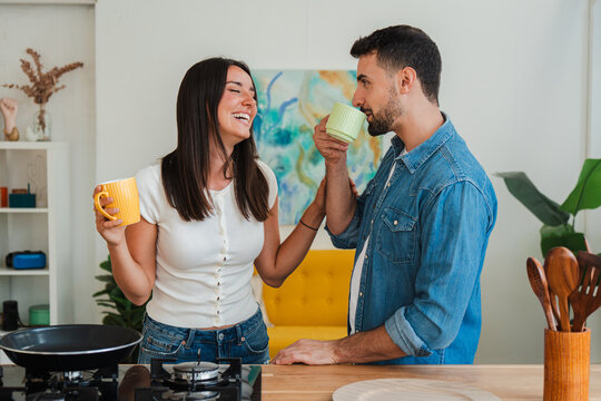 A candid shot captures a smiling couple in a sunny kitchen as they toast with matching mugs. The moment conveys warmth, connection, and shared coffee during a peaceful morning ritual.