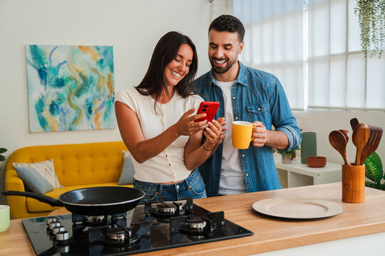 A cheerful couple browses a red smartphone while standing at a kitchen island. They hold mugs and smile, creating a warm, intimate moment of shared digital discovery during breakfast.