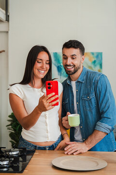 A cheerful couple photograph themselves with a bright red mobile device while standing at a wooden counter, exchanging smiles and enjoying a cozy kitchen moment as they document everyday