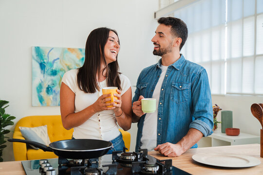 A happy couple exchange smiles while preparing breakfast together, sharing mugs in a sunny kitchen and chatting beside a stove and cookware for a warm home lifestyle moment