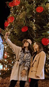 Two cheerful girlfriends taking selfie at night in front of christmas tree
