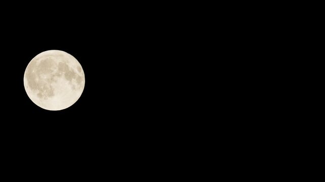 The full Moon moves across the black sky close up left side of the frame