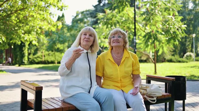 Two senior women with gray hair sitting on a park bench engaged in conversation, surrounded by greenery and walking path in a sunny outdoor setting