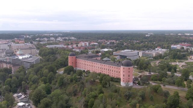 Diagonal aerial of pink Uppsala Castle on a wooded hill showing both black-domed towers and white buildings beyond.
