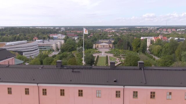 Drone along salmon-pink Uppsala Castle facade and dark roof looking over trees toward the city horizon.