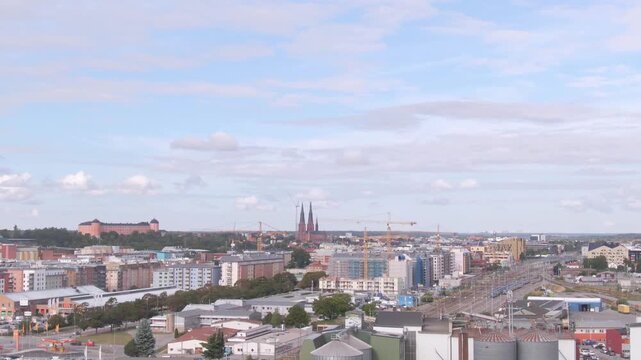 Wide aerial of Uppsala skyline under soft clouds: pink castle left, twin cathedral spires and cranes along the horizon.