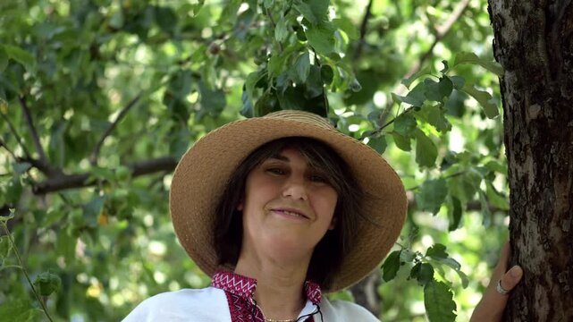 Portrait of ukrainian woman in straw hat against the background of summer garden. Adult smiling lady wearing an embroidered shirt looking into camera outdoor. National identity and patriotism concept