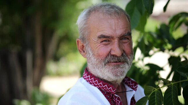 Portrait of old ukrainian man in vyshyvanka near green walnut tree of summer garden. Senior smiling male wearing an embroidered shirt looking into camera outdoor. National identity and patriotism