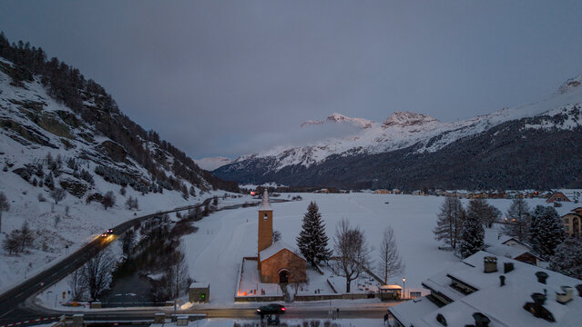 Aerial view of the historic church in Sils Maria surrounded by snow-covered mountains and a winding road at dusk in Sils im EngadinSegl, Grisons, Switzerland.