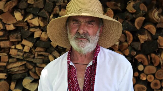 Portrait of senior ukrainian man in straw hat against the background of woodshed. Elderly serious male wearing an embroidered shirt looking into camera outdoor. Cultural heritage and ethnic roots