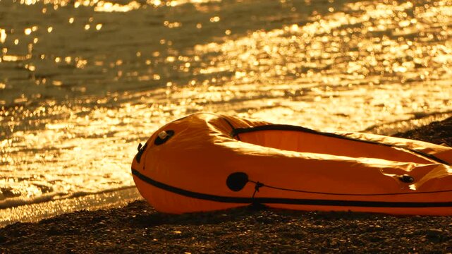 Inflatable orange dinghy rests on pebble beach shore beside golden sunlit waves at dusk. Static eye level shot.