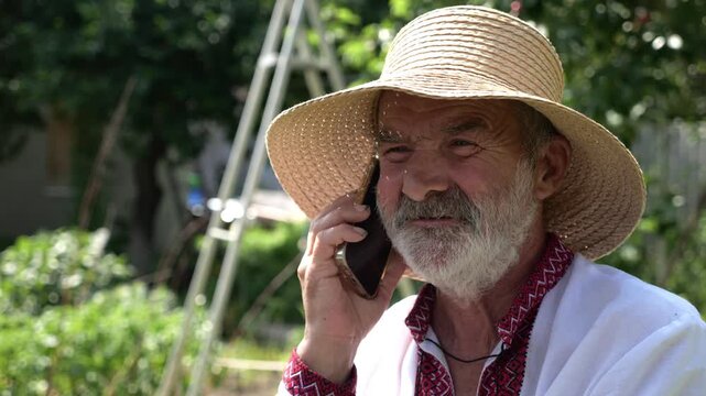Older ukrainian man wearing in vyshyvanka talking on mobile phone at summer garden. Senior male in straw hat using cell phone outdoor. Concept of connection between tradition and modern life