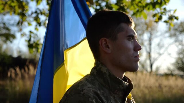 Ukrainian army man stands with national banner at countryside. Male soldier in military uniform with Ukraine flag as symbol of victory against russian aggression. Concept of invasion resistance