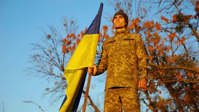 Ukrainian army man stands with national banner at countryside. Male soldier in military uniform with Ukraine flag as symbol of victory against russian aggression. Concept of invasion resistance