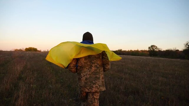Soldier of ukrainian army running with raised flag Ukraine above his head at field. Young male military in uniform jogging with blue-yellow banner at meadow. Victory against russian aggression