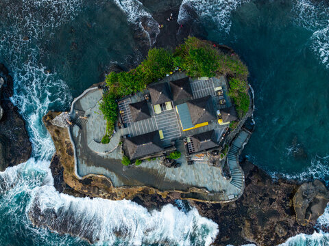 Aerial view of Tanah Lot Temple with traditional Balinese architecture and dark tiled roofs on a rocky outcrop surrounded by crashing ocean waves in Kecamatan Kediri, Bali, Indonesia.