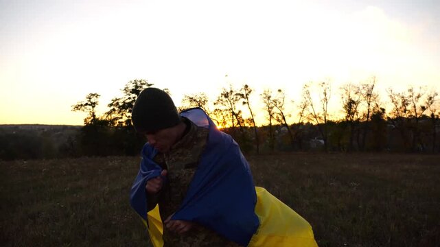 Soldier of ukrainian army going through meadow and kissing Ukraine flag as symbol of victory against russian aggression. Young male military in uniform walking with blue-yellow banner at countryside
