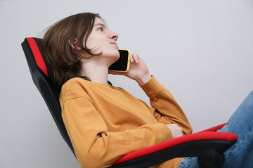 Young Person Reclining In Chair, Holding Smartphone To Ear, Wearing Mustard Sweater And Jeans, Shown In Calm Side Profile Against Plain Gray Background With Copy Space © Irina Kashparenko