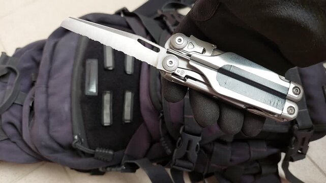 Close up top view shot of a gloved hand opening a serrated blade on a stainless steel multitool against black tactical fabric background. Concept of rescue equipment, emergency and heavy duty tools 