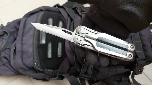 Close up top view shot of a hand in black protective work glove smoothly opening a sharp plain edge knife blade on a multitool over black tactical background. Concept of EDC tools, craft and outdoor