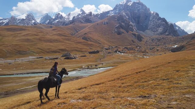 Aerial rear follow shot of traveler horseback riding across golden mountain valley towards snow-capped peaks near Kel-Suu Lake, Kyrgyzstan