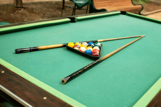 Outdoor billiard table setup with worn stained green cloth, colorful pool balls in triangle rack, selective focus cue sticks outdoors in autumn park