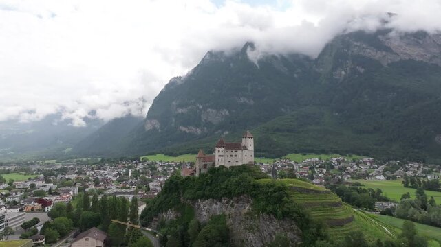 Aerial view of Vaduz Castle perched on a rocky hill above the town with lush green slopes and dramatic alpine mountains in the background, Liechtenstein.
