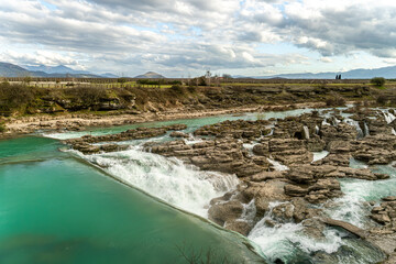 Turquoise water cascades across rugged limestone rocks on the Cijevna River in Montenegro, forming small waterfalls and rapids beneath a dramatic cloudy sky with distant Balkan mountains. © Mike Loik