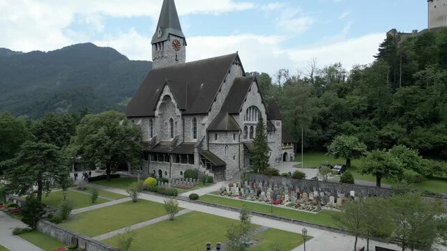 Aerial view of the historic Cathedral of St. Florin in Vaduz, featuring its stone architecture, surrounding cemetery with manicured gardens, and lush green hills in Liechtenstein.