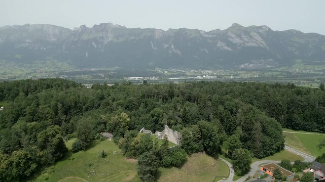 Aerial view of the historic Schalun Castle ruins surrounded by dense forest and rolling hills with the majestic Alps in the background in Liechtenstein.
