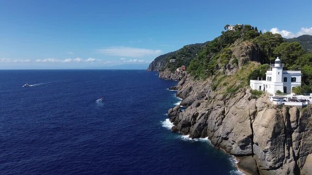 Horizontal 4K drone shot flying forward over blue waters past Portofino cliffs. Waves crash below, two boats cross the sea, white lighthouse on the cliff edge. Ungraded footage.