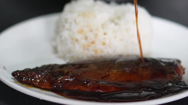 4K Sweet teriyaki sauce pouring over grilled saba fish fillet served with Japanese steamed rice on plate, representing traditional Japanese cuisine and healthy seafood menu concept.