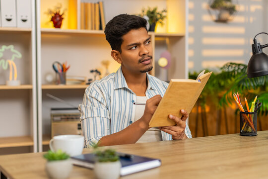 Indian man on home table reads book makes notes and pauses to think during focused study indoors at home. Arabian guy on home table reviews pages and writes ideas for learning planning and home work.
