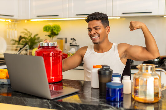 Indian man explains sports nutrition and workout supplements on laptop during meal planning at home. Arabian guy shares training advice and protein diet tips with focused face in home kitchen indoors.