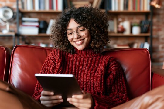 Young woman with curly hair wearing glasses sitting on a red couch using a tablet computer
