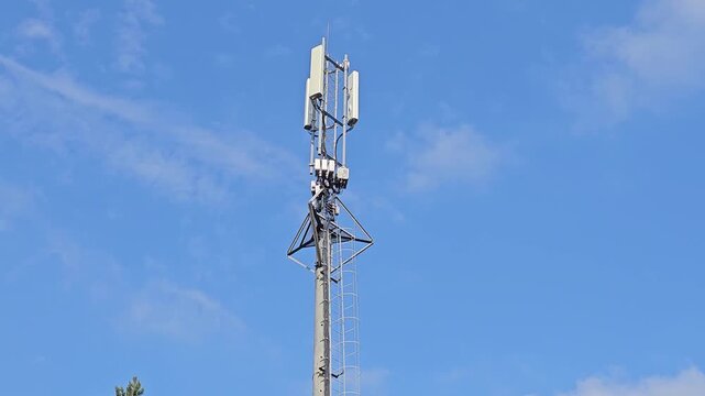 Tall tower stands against clear blue sky. White panels mounted at top catch wind and sunlight. Metal lattice supports structure with precision. Sky shows soft clouds drifting above