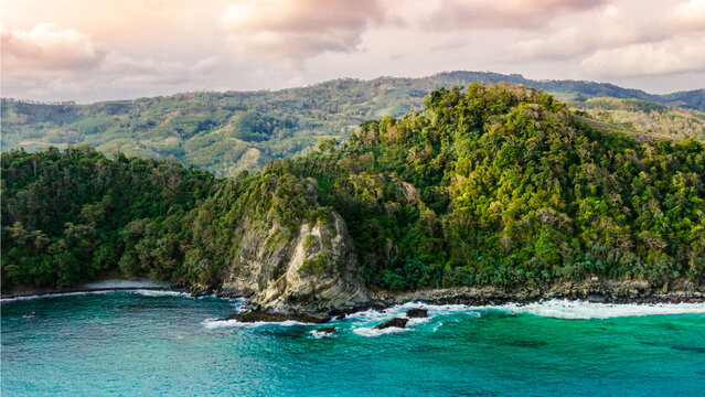 Aerial view of Modangan Beach featuring lush green cliffs, rocky outcrops, and turquoise waters under a soft sunset sky in Donomulyo, East Java, Indonesia.