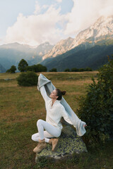 Obraz premium A woman in white clothes sits on a rock in a mountain meadow stretching her arms with a scarf. The scene shows lifestyle and relaxation in nature during daytime.