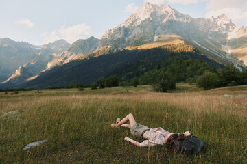 Obraz premium Young woman lies on a grassy field in casual clothes resting with a backpack near her head against a backdrop of sunlit mountains under a partly cloudy sky.