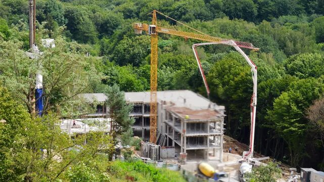 A construction site in a green forest features a yellow tower crane and a concrete pump truck with a hydraulic boom pouring liquid concrete onto the building frame and a drilling rig.