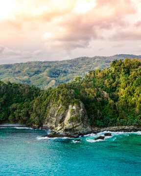 Aerial view of Modangan Beach featuring lush green hills meeting the turquoise sea with rocky cliffs and soft sunset light in Donomulyo, East Java, Indonesia.