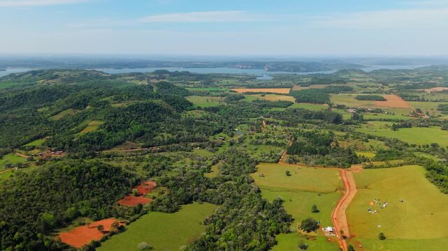 Sweeping drone panorama of Nueva Alborada, Paraguay. Lush green hills, agricultural fields, and vibrant red dirt roads stretch toward a distant vast river.
