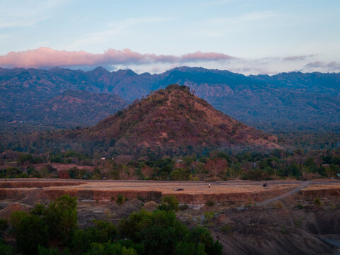 Aerial view of Savana Tianyar with its prominent hill, dry grassy plains, and distant mountains under a soft sunset sky in Kecamatan Kubu, Bali, Indonesia.