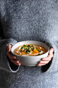 Female hands in gray knitted sweater holding bowl of pumpkin soup