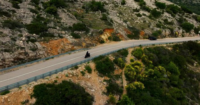 A drone follows a motorcyclist along a winding coastal road at Stara Baska on Krk island, Croatia. Rocky hills and green shrubs rise above blue water.