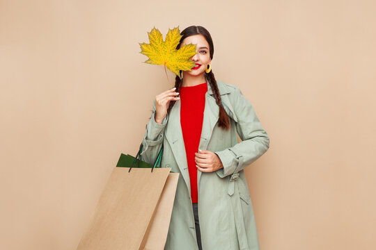 Autumn portrait of beautiful happy young woman with shopping bag, stylish girl holding yellow maple leaves in round hat, coat on beige studio background