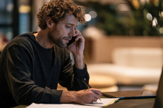 A focused man engages in a phone call while jotting down notes at a desk, highlighting themes of productivity, communication, and modern workspaces. The cozy environment enhances the moment.
