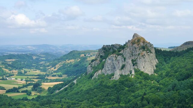 panoramic view of rocky mountain peak surrounded by green forest and rural valley under cloudy sky in auvergne france