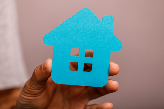A person's hand gently holds a vibrant blue paper house, representing real estate and dreams.